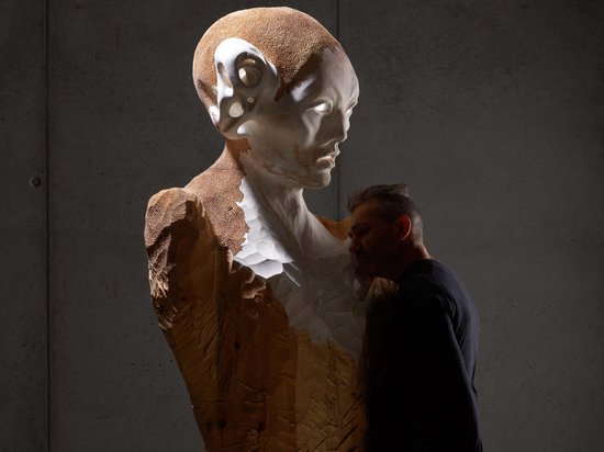Sensoria Art Edition 01 Mann betrachtet eine große hölzerne Büste mit detailliertem Gesicht gegen Betonhaut