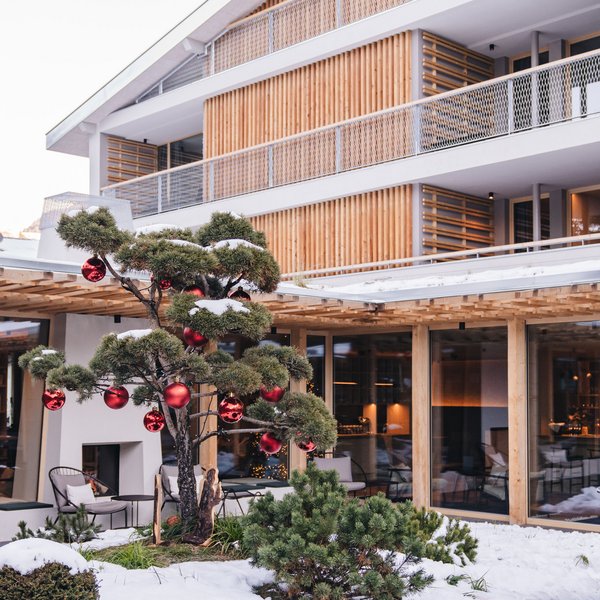 Sensoria Dolomites: Wellnesshotel in den Dolomiten Moderner Holzbau mit verschneitem Garten und geschmücktem Baum mit roten Kugeln