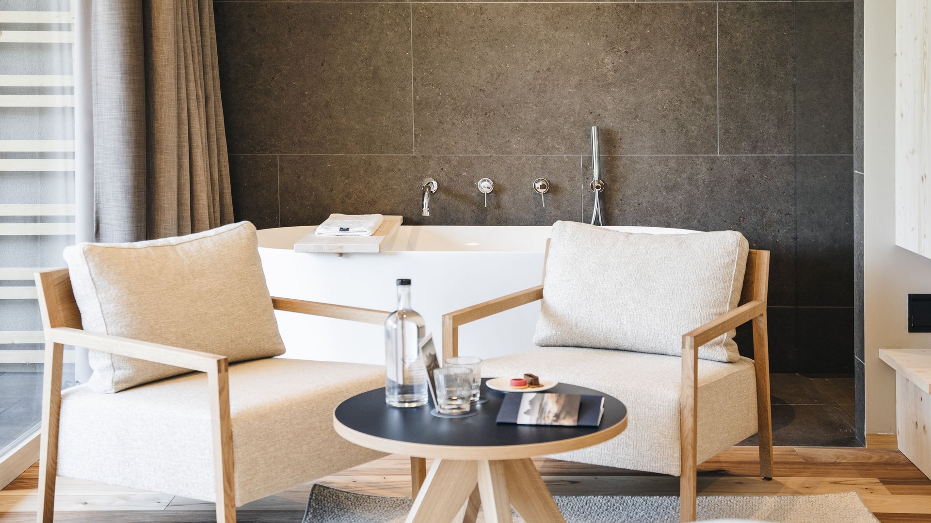 Inclusive services – last-minute South Tyrol holidays Two beige armchairs and table in front of bathtub with dark tile wall
