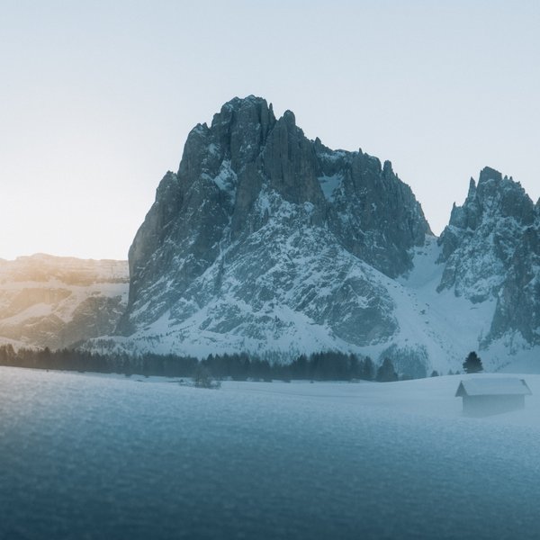 Aktivurlaub in Südtirol – bereit für Natur pur? Schneebedeckter Berg bei Sonnenaufgang mit Hütte und Bäumen im Vordergrund