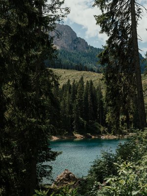 Luxus für die Seele im Kurzurlaub in Südtirol Bergsee mit klar blauem Wasser umgeben von Tannen und Bergen bei bewölktem Himmel