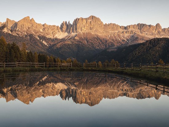 Die Dolomiten – Skiurlaub und so viel mehr Berglandschaft mit Spiegelung in stillem Wasser und Holzzaun im Vordergrund