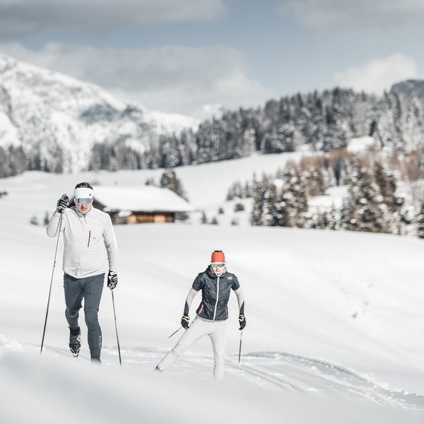 Seiser Alm: Skiurlaub de luxe Zwei Menschen beim Skilanglauf in verschneiter Berglandschaft mit Hütte