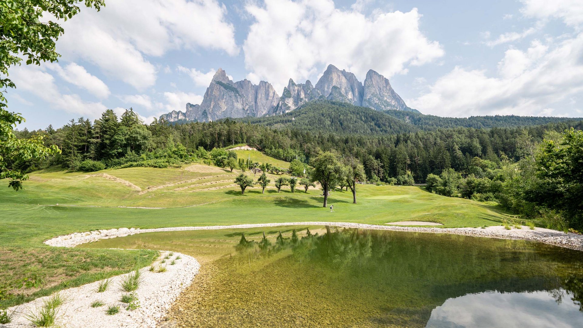 Golf und Wellness – Ihr Golfhotel nahe der Seiser Alm Berglandschaft mit See, Bäumen und grünen Wiesen unter bewölktem Himmel