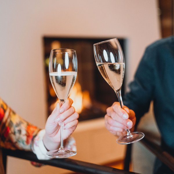Genussurlaub in Südtirol – Sensoria Dolomites Zwei Personen stoßen mit Sektgläsern vor einem Kamin an