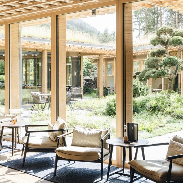 Nachhaltiges Handeln im Sensoria Dolomites Helles Loungezimmer mit großen Fenstern und Blick auf einen Garten mit Bäumen
