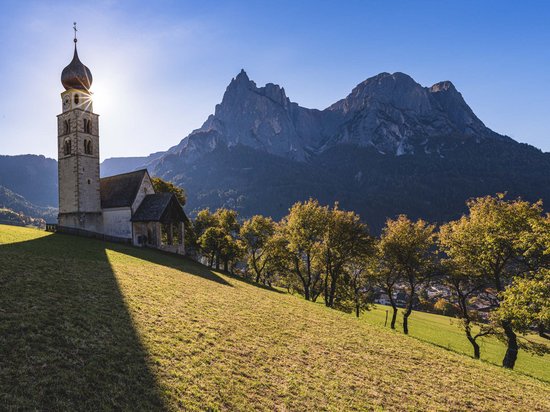 Die Dolomiten – Skiurlaub und so viel mehr Kirchturm in Berglandschaft mit Bäumen und Sonne im Hintergrund