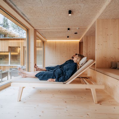 Naturhotel in Südtirol. Luxus für die Seele. Paar entspannt in blauen Bademänteln auf Holzliegen in modernem Spa mit Fensterblick