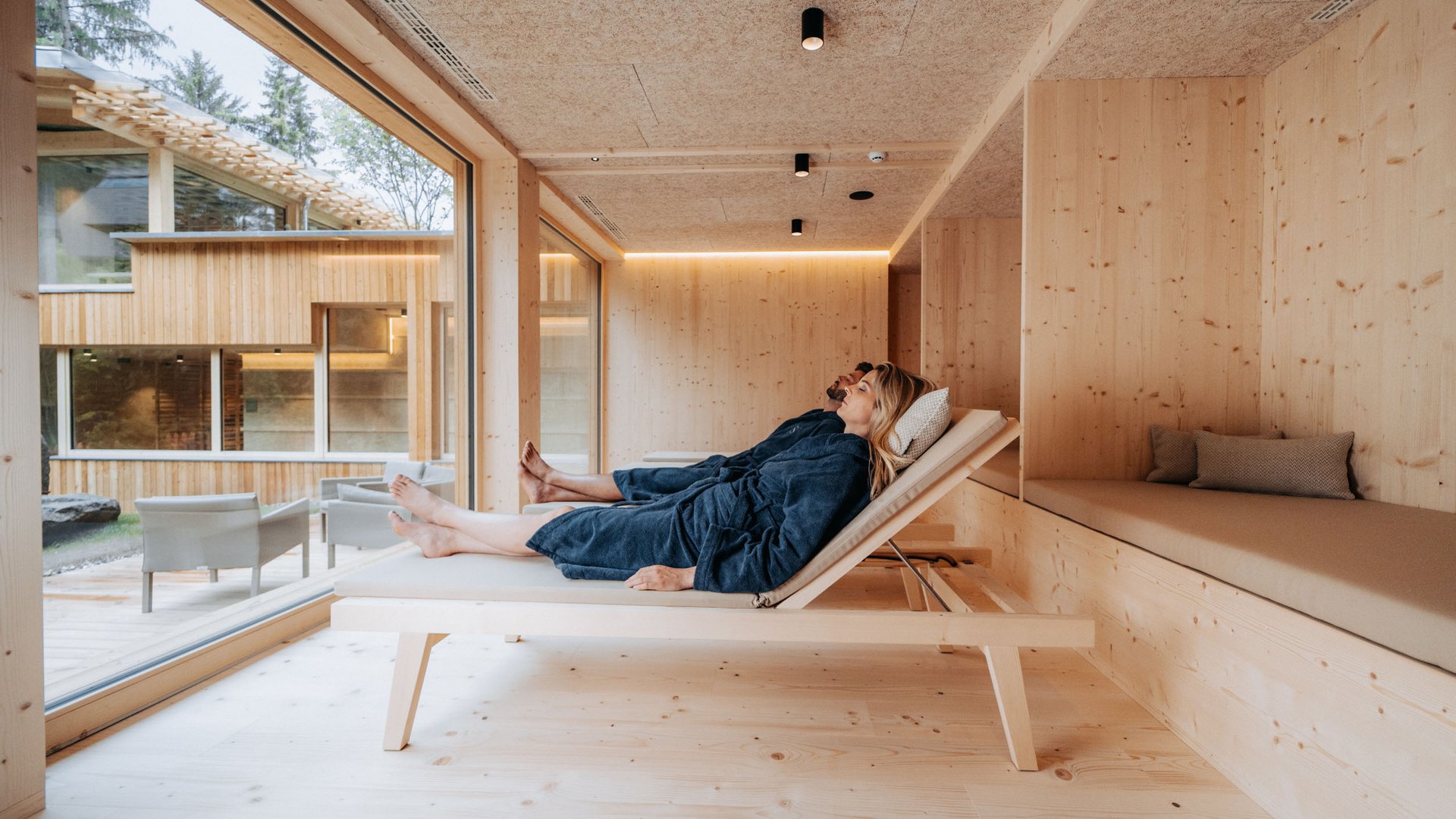 Von Wanderungen auf der Seiser Alm in die Spa Area Paar entspannt in blauen Bademänteln auf Holzliegen in modernem Spa mit Fensterblick