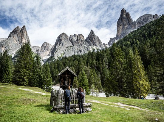 Die Dolomiten – Skiurlaub und so viel mehr Wanderer betrachten Kreuzigungsfigur in den Dolomiten vor Nadelwald und Bergen