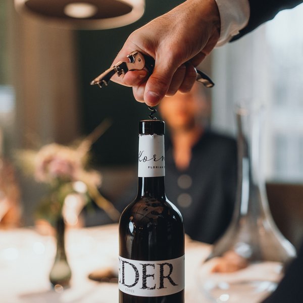 Genussurlaub in Südtirol – Sensoria Dolomites Hand öffnet Weinflasche mit Korkenzieher an gedecktem Tisch