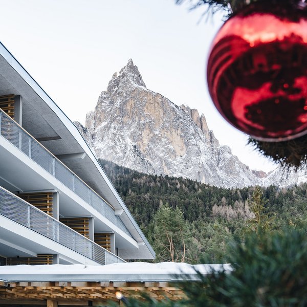 Sensoria Dolomites: Wellnesshotel in den Dolomiten Schneebedeckter Berg hinter einem modernen Gebäude und Weihnachtsbaum mit roter Kugel