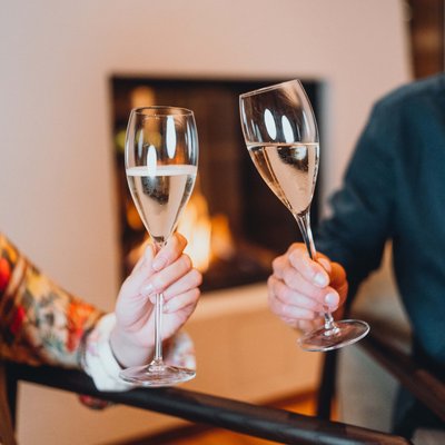 Essence West Zwei Personen stoßen mit Sektgläsern vor Kamin an