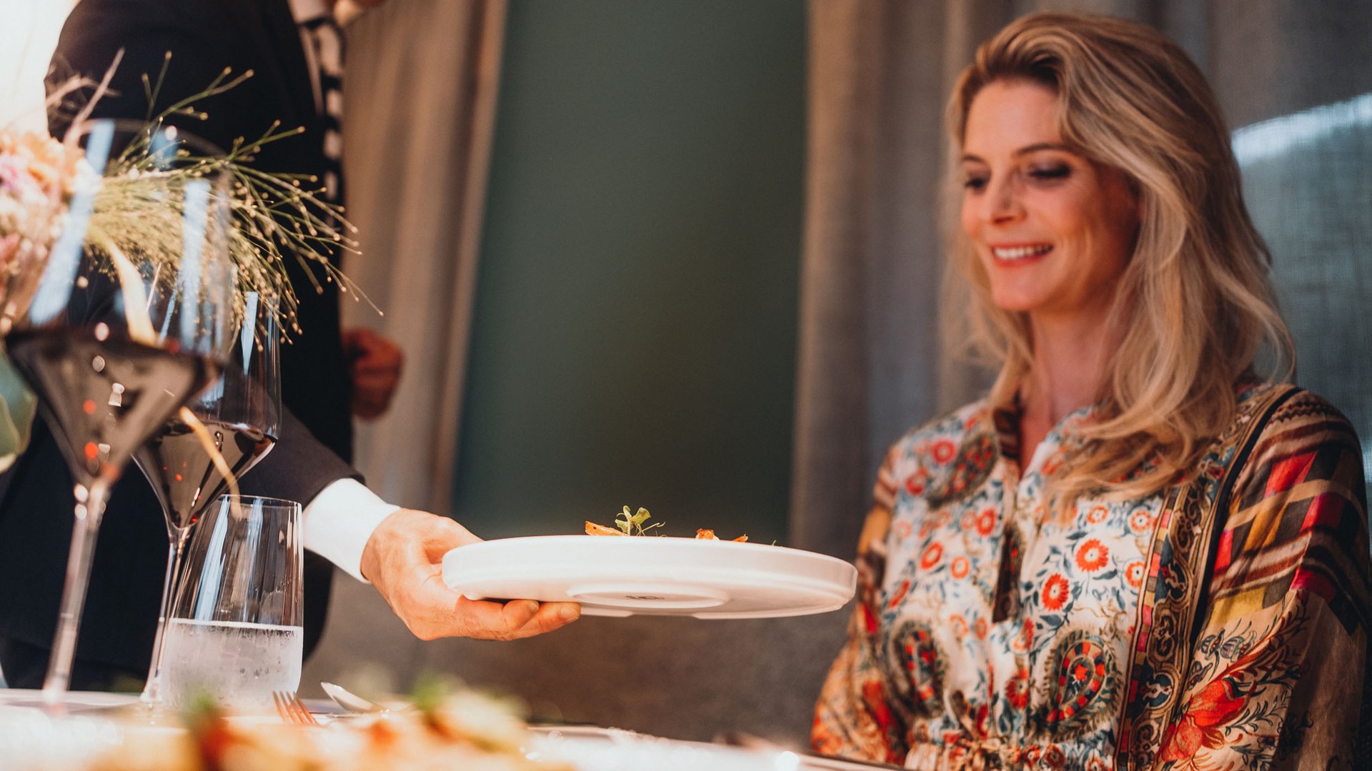 Genussurlaub in Südtirol – Sensoria Dolomites Kellner serviert einem lächelnden Frau in gemustertem Kleid ein Gericht