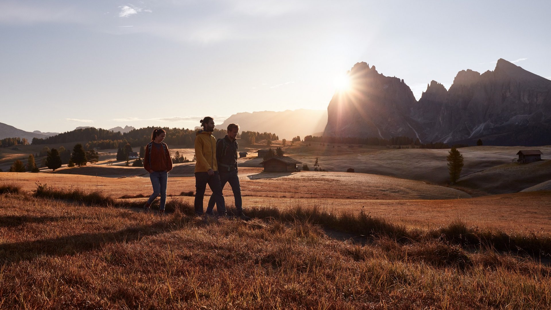 Mehr als ein Hotel auf der Seiser Alm: Sensoria Dolomites Drei Wanderer gehen bei Sonnenuntergang durch eine herbstliche Landschaft mit Bergen