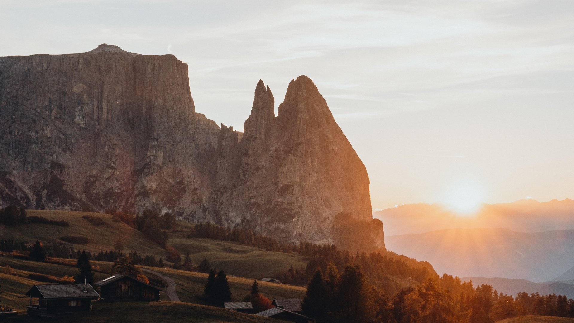Hotel in Seis am Schlern gesucht? Sensoria Dolomites gefunden! Sonnenuntergang hinter Felsspitzen in einer bergigen Landschaft mit Hütten