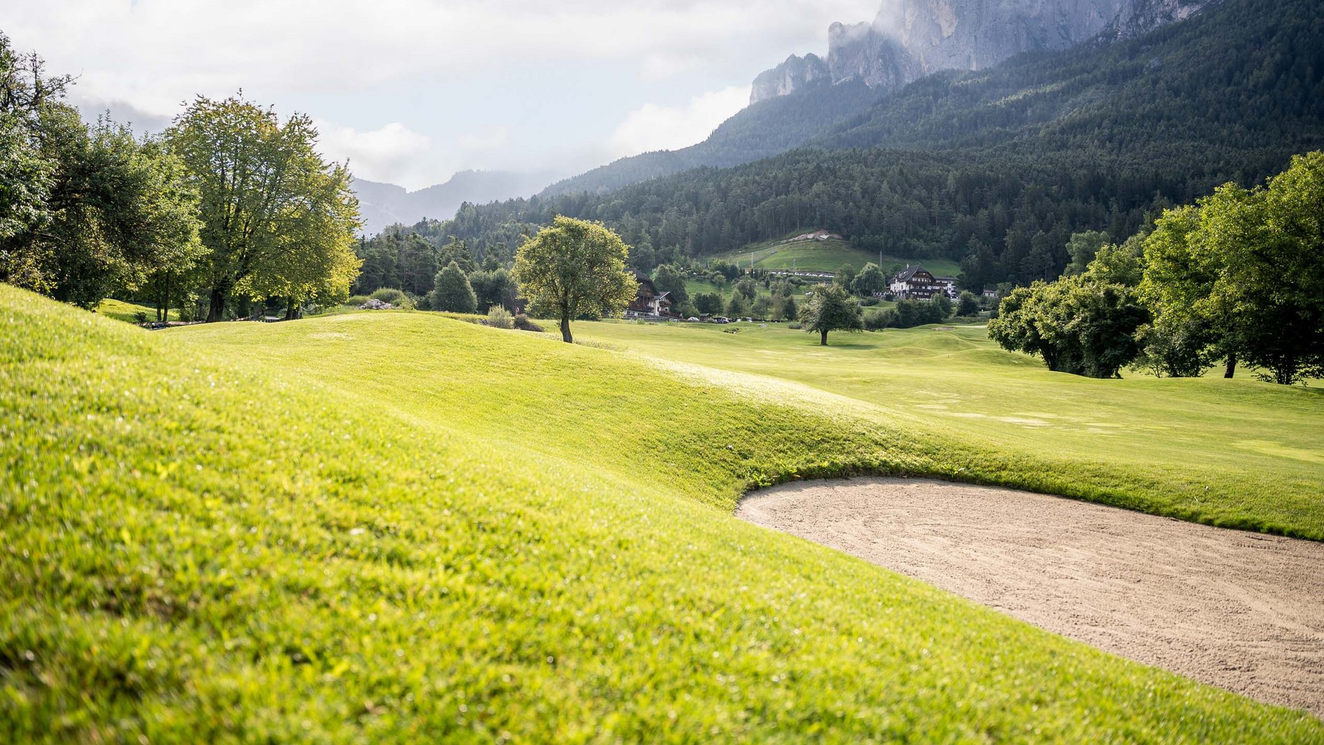 Golf und Wellness – Ihr Golfhotel nahe der Seiser Alm Grünes Golfplatzgelände mit Bunker und Bergen im Hintergrund