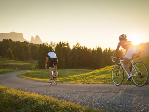Ein Sommerurlaub in den Bergen Zwei Radfahrer fahren bei Sonnenuntergang auf einer Landstraße durch grüne Landschaft.