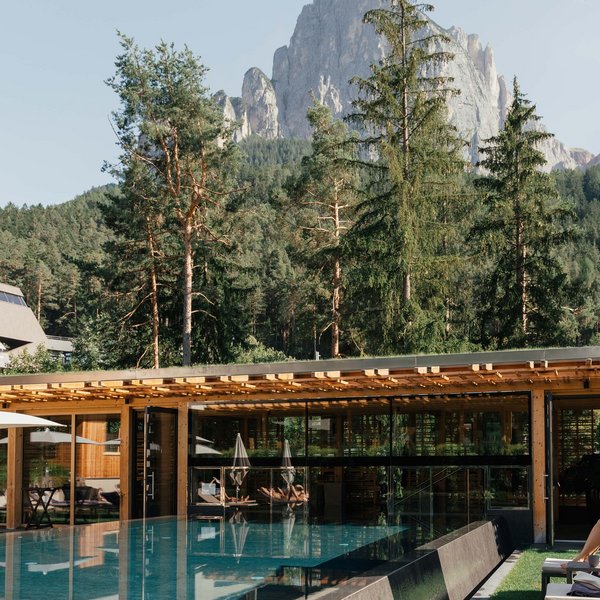 Von Wanderungen auf der Seiser Alm in die Spa Area Moderner Pool mit gläsernem Gebäude vor bewaldetem Berg in sonniger Landschaft
