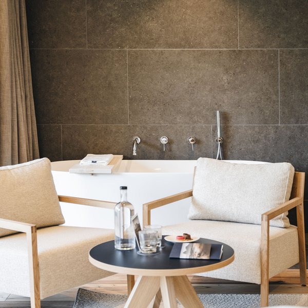 Your hotel in the Dolomites: Sensoria Dolomites Two beige armchairs and table in front of bathtub with dark tile wall