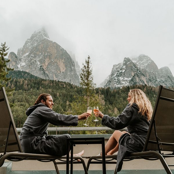 Your hotel in the Dolomites: Sensoria Dolomites Couple in robes toasting drinks on terrace with mountain view
