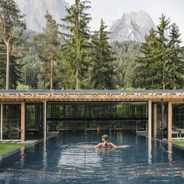 Your hotel in the Dolomites: Sensoria Dolomites Woman in pool with view of forested mountains and modern building